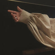 grainy photo of a hand reaching out to the left of the screen. the hand is slightly closed and the arm is clothed in white linen