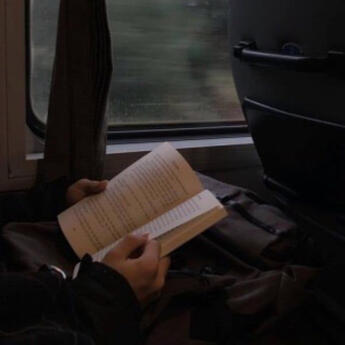 focuses on an open book being held by the passenger of a train. in the background, a rainy scene can be seen racing passedl